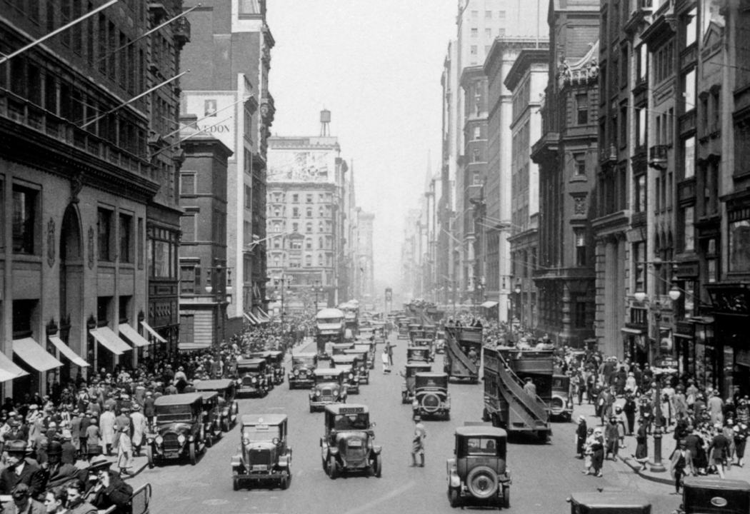 Imagen histórica en blanco y negro de una bulliciosa calle de la ciudad, poblada de peatones y coches.